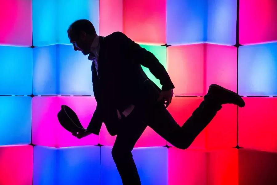 Image of man in front of glowing boxes, posing and lowering hat in a bow position